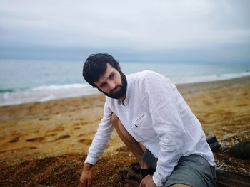 Bearded young man sitting at beach against cloudy sky