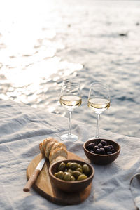 Two glasses of white wine and a wooden plate with bread and olives on a pier at sunset.