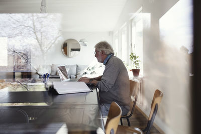 Senior man using laptop while sitting at table seen through window