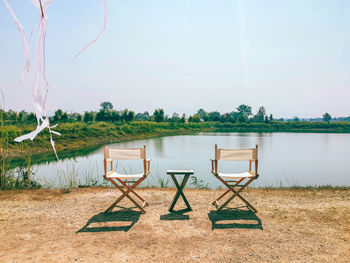 Empty chairs by lake against sky