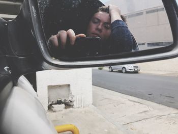 Reflection of man taking selfie through mobile phone on side-view mirror of car