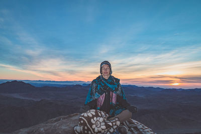 Woman meditating while sitting on rock against sky during sunset