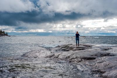 Rear view of mature woman standing on rock at beach against cloudy sky