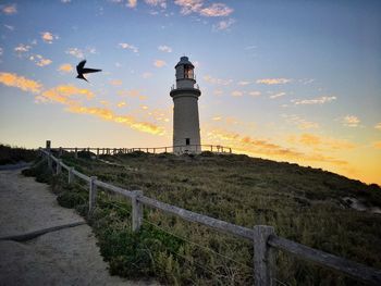 Bird flying by lighthouse against sky during sunset