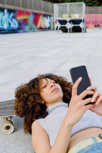 High angle of cheerful black woman with afro hair lying on skateboard on paved ground and using phone to take selfie in town