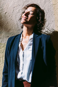 Thoughtful happy female wearing elegant clothes standing sunlight and casting shadow on shabby wall with eyes closed