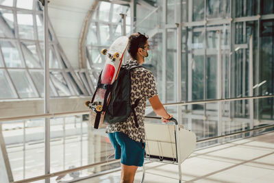 Young man with face mask walking at the airport with skateboard on his back