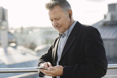 Mature businessman listening music through phone while standing at office