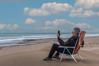 Mature adult male sitting on a beach chair on dune facing the sea talking on video with smartphone.