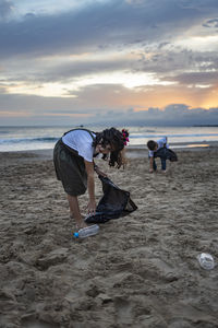 Girl and boy collecting garbage at beach against sky during sunset