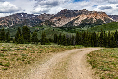 Dirt road by trees and mountains against sky