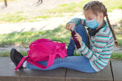 A cute caucasian girl in a mask sits in the schoolyard with a backpack and a phone in her hands.