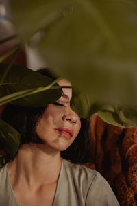 Through leaves of green plant of delicate female sitting in leather armchair at home and closed eyes