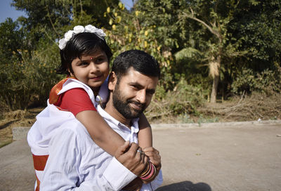 Portrait of smiling man piggybacking daughter against trees