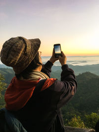 Woman photographing nature while standing against sky during sunset