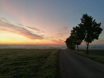 Empty road amidst silhouette trees on field against sky at sunset