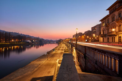 Illuminated buildings by river against sky at sunset