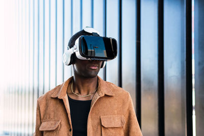 African american male in vr goggles exploring cyberspace while standing on street near modern building with metal wall in city