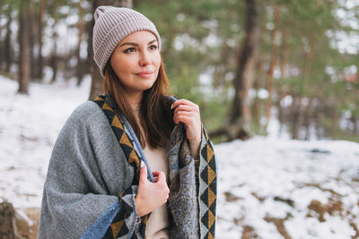 Portrait of young brunette beautiful woman in hat and grey poncho in the winter forest