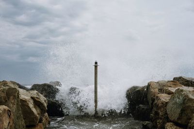 Waves splashing on pole at shore against cloudy sky