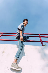 From below side view of of brave male hanging looking away over metal fence in street and showing parkour trick against blue sky