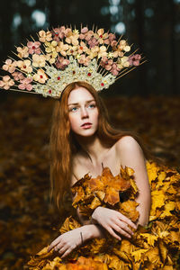 Thoughtful young woman covered with leaves wearing flowers at forest during autumn