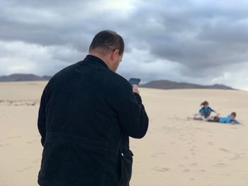 Rear view of man using mobile phone while standing at beach against cloudy sky