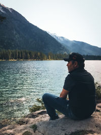 Side view of young man sitting on lake against mountains