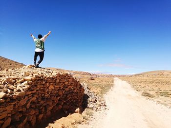 Rear view of man with arms outstretched standing on stone wall by dirt road at desert against blue sky