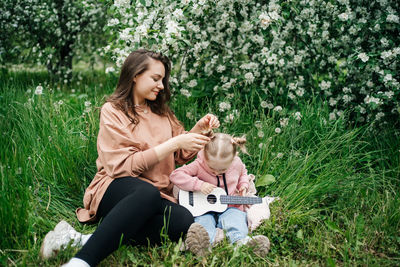Young mother and baby daughter play the ukulele in a blooming apple orchard in nature