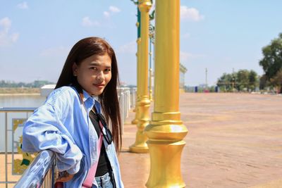 Portrait of smiling woman standing by pole against sky