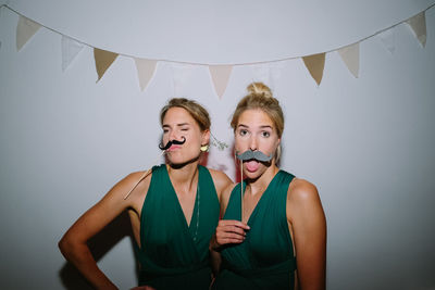 Portrait of young woman with twin sister holding mustache props against wall