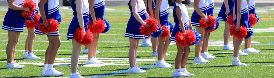 Young cheerleaders in white and blue uniforms holding red and blue pompoms on a turf field.