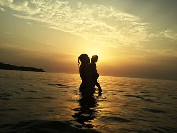 Silhouette mother with son standing in sea against sky during sunset