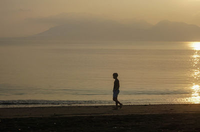 Rear view of woman walking at beach against sky during sunset