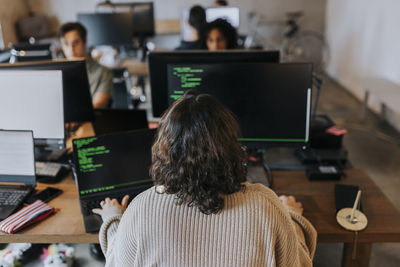 Rear view of young female programmer coding on computer at desk in office