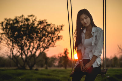 Beautiful young woman sitting on swing against sky during sunset