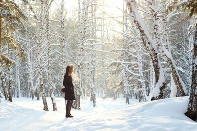 Side view of woman standing against bare trees on snow covered field at forest