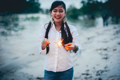 Cheerful young woman holding illuminated sparklers by lake against sky