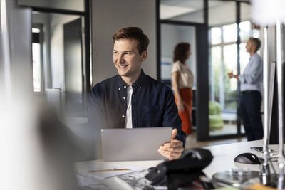 Smiling businessman looking away while sitting with colleagues in background at open plan office