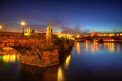 Illuminated bridge over river by buildings against sky at night