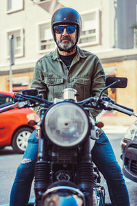 Serious bearded man in sunglasses and helmet sitting on motorbike during ride on city street looking at camera