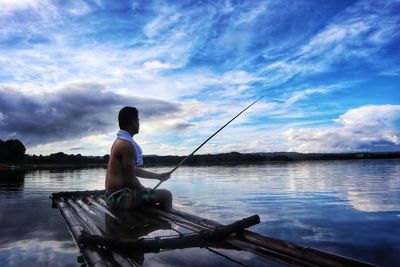 Shirtless man fishing while sitting on raft against sky