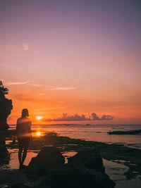 Rear view of man standing on rock by sea against sky during sunset