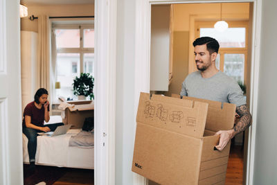 Smiling man carrying box from doorway towards woman using laptop in bedroom