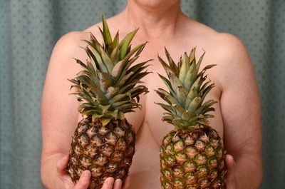 Midsection of shirtless mature woman holding pineapples by curtain