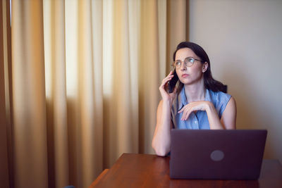Woman with glasses a freelancer with a laptop sitting at a table in apartment. talking on the phone