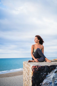 Young woman sitting on rock at beach against sky