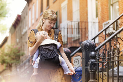 Mother holding baby in carrier while walking against houses