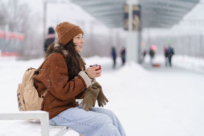 Tired but happy female traveler with mug of hot tea waiting for train to arrive in wintertime